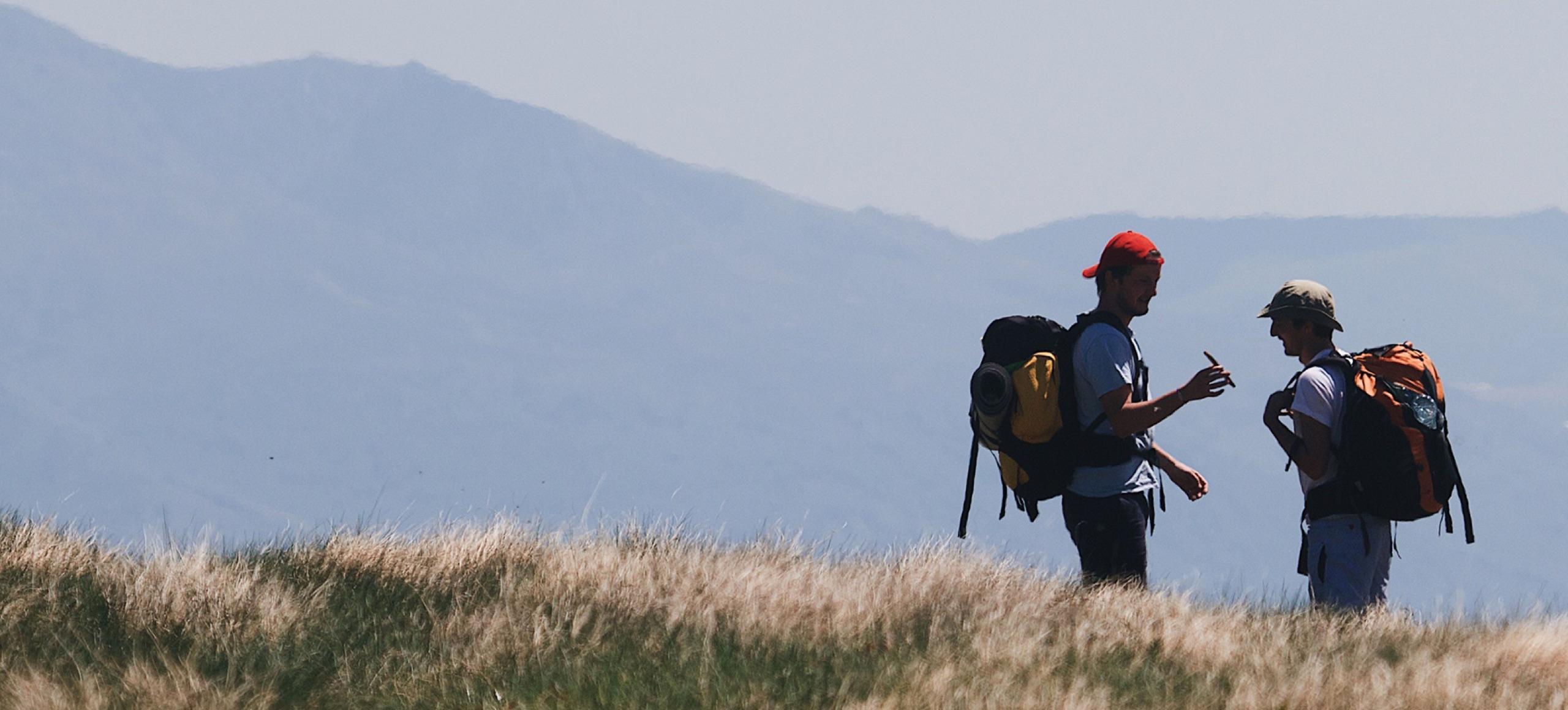 Dos viajeros con mochila conversando en una pradera de montaña