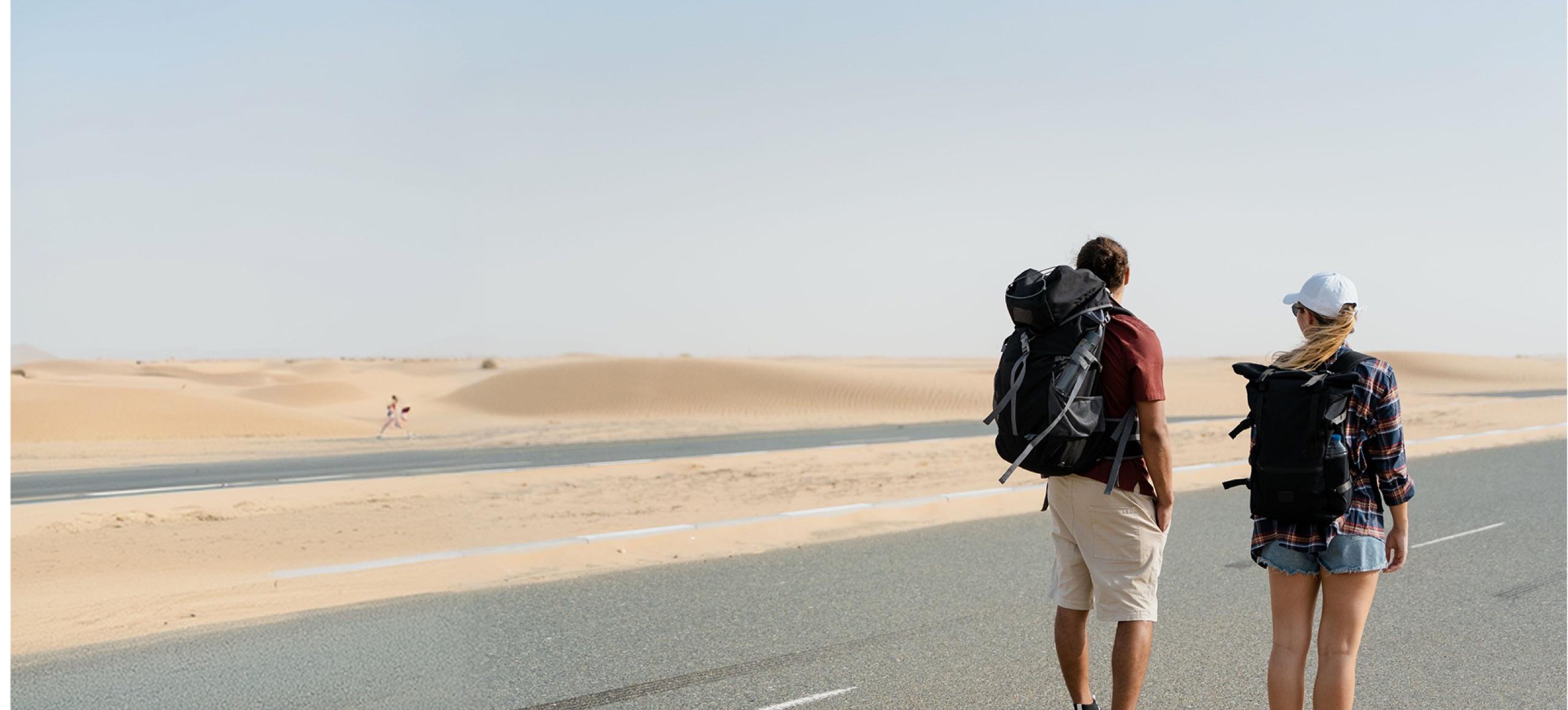 Dos viajeros con mochila caminando por una carretera entre dunas del desierto